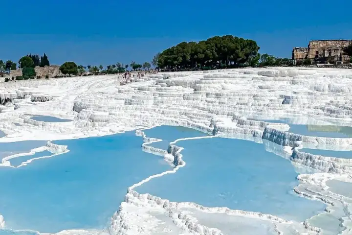 Pamukkale's stepped travertine terraces in Turkey under a bright blue sky, wide turquoise thermal pools separated by flat white calcium ridges descending in orderly tiers, tourists visible walking the upper terrace as pine trees and ancient Hierapolis ruins appear at the top.