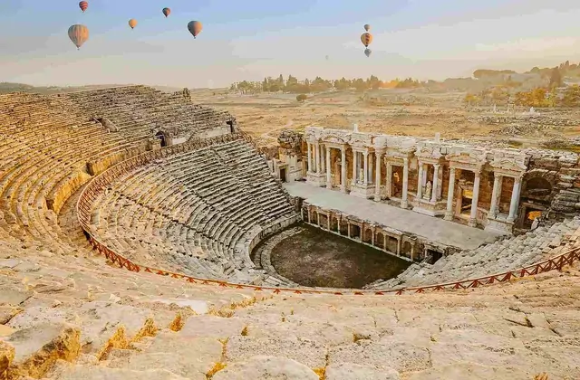 The Hierapolis theater at Pamukkale seen from the upper seating tiers at golden hour with hot-air balloons floating overhead, the ornate Roman stage building glowing warm amber as the vast Denizli plain stretches to distant mountains under a hazy horizon.