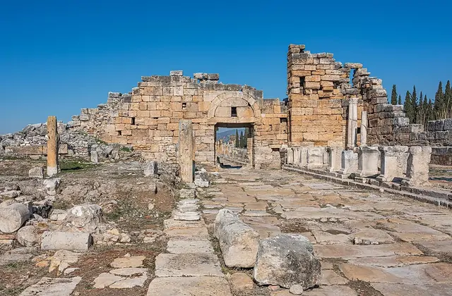 The Domitian Gate and Frontinus Street ruins of Hierapolis at Pamukkale, Turkey, viewed from the wide cobblestone road looking toward the partially collapsed stone archway, fallen column drums lying on the ground and cypress trees visible through the arch.