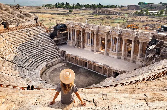 A female tourist wearing a wide sun hat sits on the stone seating of the Hierapolis theater at Pamukkale, Turkey looking out over the ancient Roman stage with ornate columned facade, the surrounding excavation site and valley visible beyond.