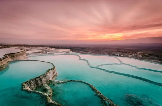Aerial view of Pamukkale's white travertine terraces with vivid teal-green serpentine pools at pink-orange sunset in Turkey, the large sinuous basins glowing under a dramatic rose sky while the distant Denizli valley fades toward the horizon.