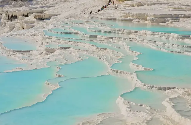 Mid-distance view of Pamukkale's stepped travertine terraces in Turkey showing visitors walking along the upper white ridge and dozens of wide turquoise pools in an organic cellular pattern, icicle-like calcium stalactites hanging from terrace edges.