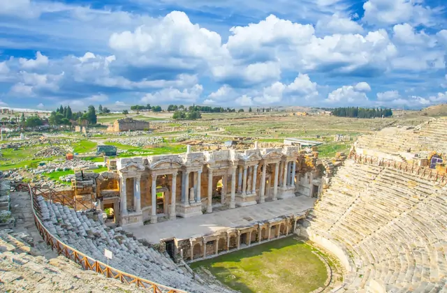 Pamukkale's Cotton Castle terraces and turquoise pools viewed from above in Turkey under a bright blue winter sky with wisps of cloud, icy-white calcium carbonate ledges descending in steps toward the wide valley below, the town visible at the base of the hill.