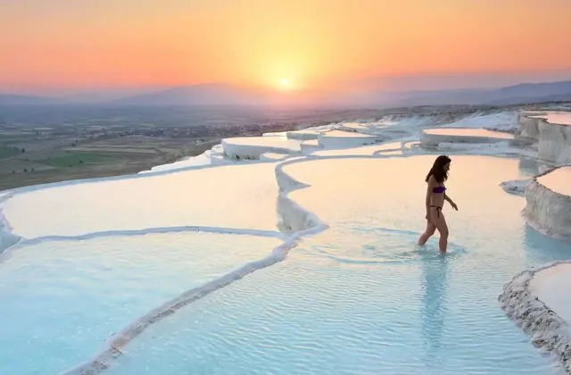 Pamukkale's white travertine terraces and turquoise thermal pools cascading down the Cotton Castle hillside in Turkey, a wide landscape view showing the layered calcium formations against a vivid blue sky with mountains in the background.