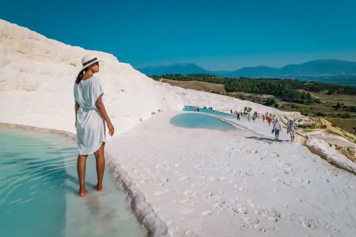 A woman in a white short dress and sun hat wades barefoot along a Pamukkale travertine terrace pool edge in Turkey, groups of tourists visible further along the white hillside and the turquoise lower pool catching sunlight under mountains.
