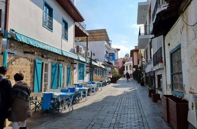 Una strada acciottolata nel Kaleici di Antalya fiancheggiata da edifici intonacati di bianco e rivestiti in pietra con telai di finestre e persiane in legno nei toni del verde acqua e turchese, tavolini da caffè all'aperto disposti sul marciapiede sinistro sotto un cielo invernale terso.
