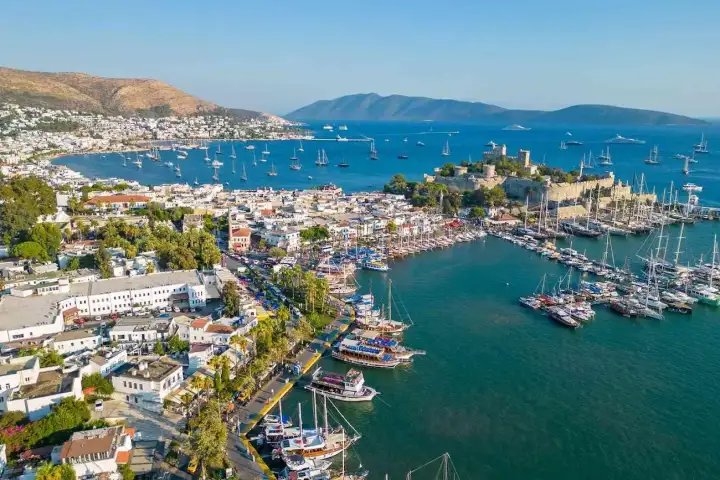 Una toma aérea de dron mirando hacia el frente marítimo del centro de Bodrum captura el concurrido paseo marítimo del puerto bordeado de árboles y edificios coloridos a orillas del agua, docenas de gulets y veleros amarrados a lo largo del muelle, y el Castillo de San Pedro de Bodrum visible a media
