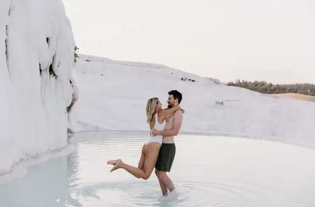 A couple embraces romantically in a shallow Pamukkale travertine pool in Turkey, the woman in a white swimsuit lifted by the man, the towering snow-white travertine cliff wall filling the background and distant green forest just visible at the top.