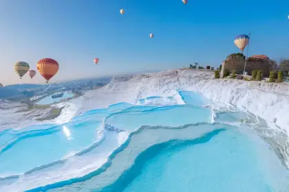 Colorful hot-air balloons float over Pamukkale's vivid turquoise travertine terraces on a bright morning in Turkey, the ancient Byzantine tower ruin visible on the upper ridge among flowering bushes as balloons reflect in the pool water below.