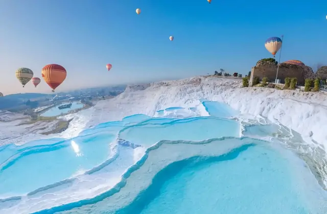 Colorful hot-air balloons float over Pamukkale's vivid turquoise travertine terraces on a bright morning in Turkey, the ancient Byzantine tower ruin visible on the upper ridge among flowering bushes as balloons reflect in the pool water below.