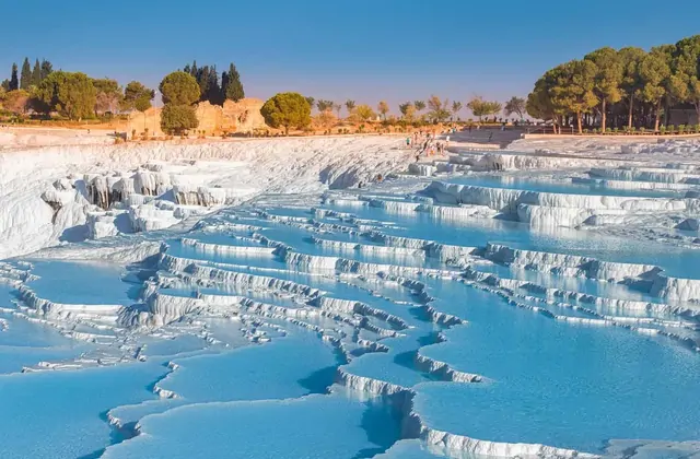 Pamukkale's white travertine terraces filled with vivid blue thermal pools and dense rows of parallel narrow stepped ledges under warm afternoon light in Turkey, Hierapolis ruins and umbrella pine trees visible atop the upper plateau.