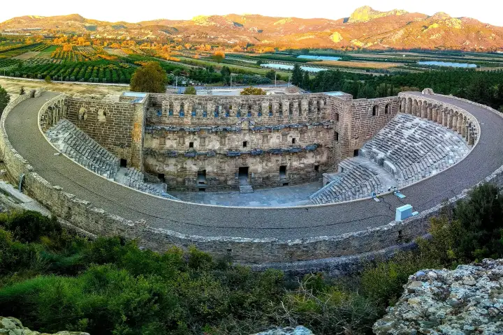 An elevated aerial view of the Aspendos Roman theater in Antalya, Turkey, reveals the complete horseshoe-shaped cavea and its towering two-storey stage building, with the fertile Pamphylian plain and mountain ridges stretching to the horizon.