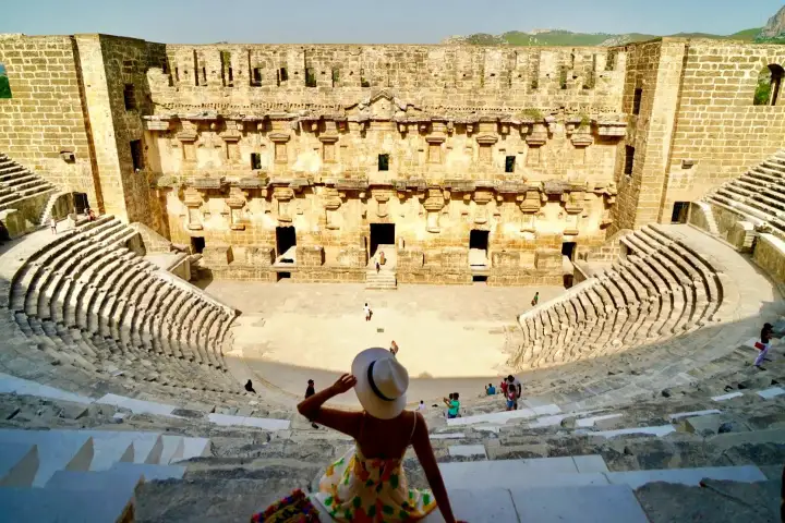 Una visitante solitaria con vestido floral se sienta en los escalones de piedra del teatro de Aspendos en Antalya, Turquía, contemplando las gradas semicirculares hacia el imponente muro escénico, con sus múltiples puertas y piedras erosionadas enmarcadas por un cielo azul despejado.