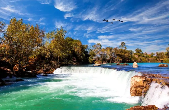 La Cascata di Manavgat vicino a Side, Antalya, precipita su un'ampia cornice naturale in una pozza di brillante acqua turchese-verde, con lussureggianti alberi dalle sfumature autunnali che costeggiano entrambe le rive e uno stormo di uccelli che vola su un vivace cielo azzurro con nuvole bianche.