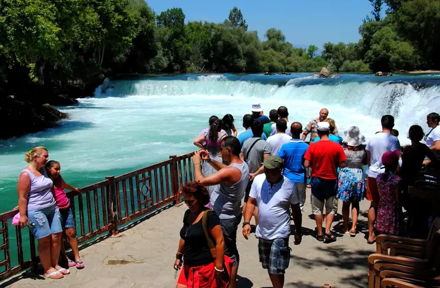 Una multitud de turistas se apoya en la barandilla de un paseo fluvial en la Cascada de Manavgat, cerca de Side, Antalya, mientras el río se precipita sobre una amplia cascada hacia las espumosas aguas turquesas bajo un brillante cielo veraniego.
