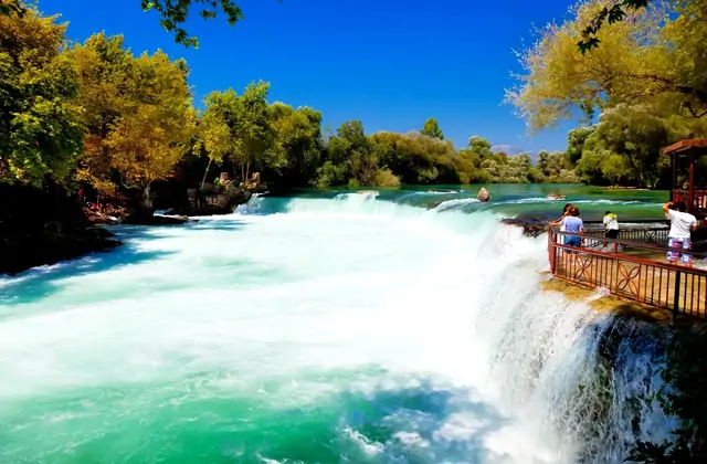 Dos visitantes están de pie en una plataforma de madera en voladizo al borde de la Cascada de Manavgat cerca de Side, Antalya, observando el ancho río precipitarse sobre las cascadas en un torrente de espuma blanca hacia la cristalina poza turquesa de abajo, rodeada de árboles verdes bajo un cielo a