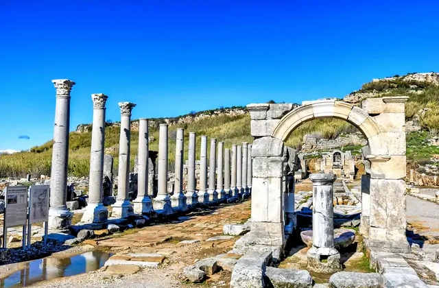 Una lunga fila di alte colonne corinzie in marmo costeggia l'avenue colonnata dell'antica città di Perge vicino ad Antalya, Turchia, conducendo verso un arco in pietra con la collina dell'acropoli ricoperta di erba che si erge sullo sfondo sotto un brillante cielo azzurro.