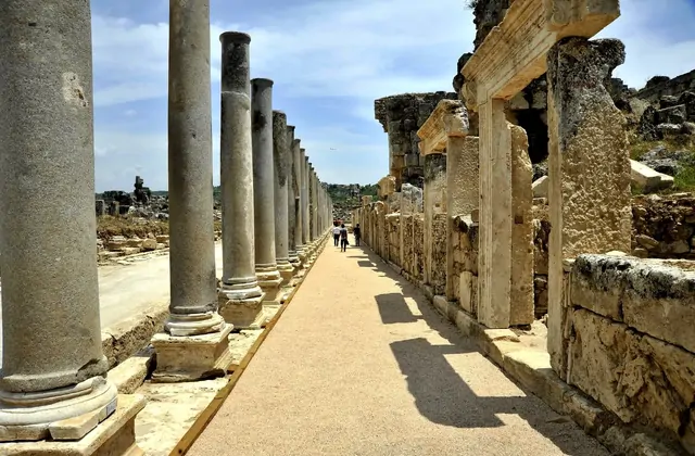 Dos figuras distantes caminan por el sendero peatonal central de la calle columnata principal de Perge, Antalya, flanqueadas por altas columnas de granito a un lado y un muro continuo de sillería con puertas talladas al otro, que se aleja hacia el horizonte bajo un cielo parcialmente nublado.