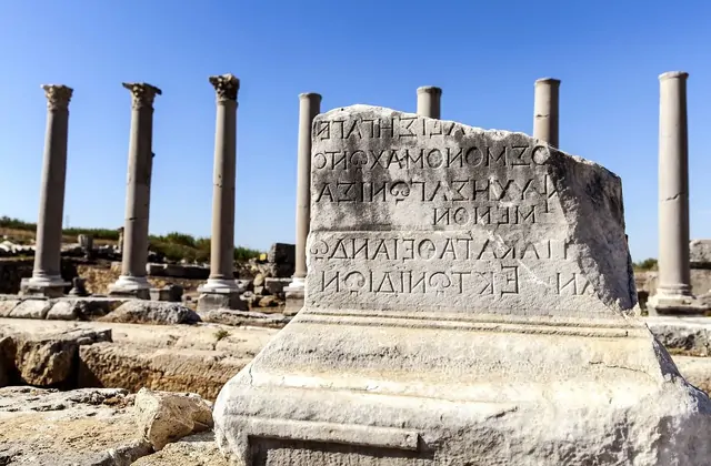 A close-up of an ancient marble inscription block at Perge, Antalya, Turkey, shows multiple lines of incised Greek lettering in sharp relief, with a row of Corinthian column shafts and blue sky visible in the background.