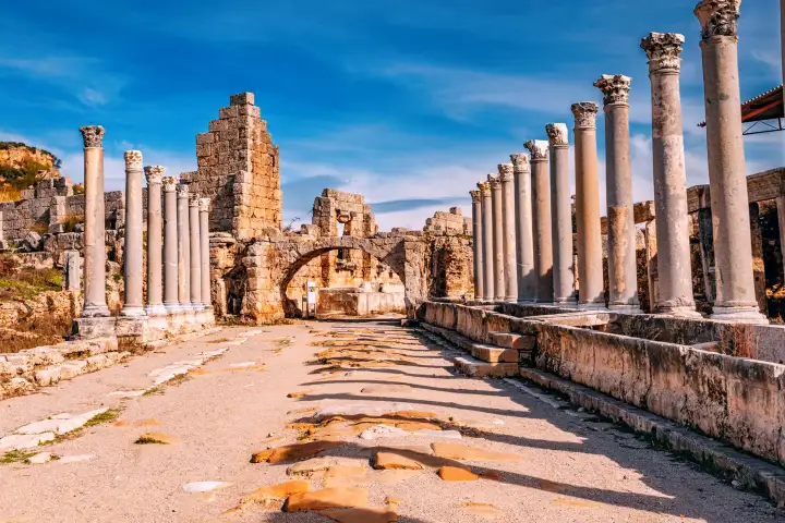 La monumental puerta helenística y las torres redondas que flanquean la entrada a la muralla de la antigua ciudad de Perge, Antalya, Turquía, se elevan sobre el patio abierto pavimentado con piedras antiguas, con sus macizos muros de mampostería con nichos abovedados alzándose contra un cielo azul b