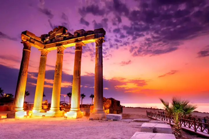 Four tall Corinthian columns of the Temple of Apollo at Side, Antalya, are dramatically illuminated against a blazing magenta and violet sunset sky, their warm golden-lit shafts and partially intact entablature silhouetted above the Mediterranean shoreline.