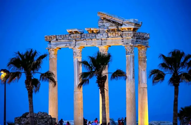Il Tempio di Apollo a Side, Antalya, brilla sotto una calda illuminazione artificiale su un profondo cielo azzurro serale, con le sue quattro colonne corinzie in piedi e i frammenti di fregio scolpito incorniciati da palme in silhouette mentre i visitatori si radunano alla sua base.