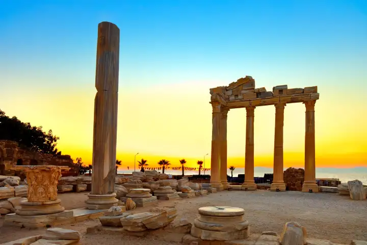 At twilight in Side, Antalya, a single isolated column and the four-column portico of the Temple of Apollo stand side by side in golden sunset light above a field of scattered marble architectural fragments, with the calm Mediterranean Sea glowing on the horizon beyond palm trees.