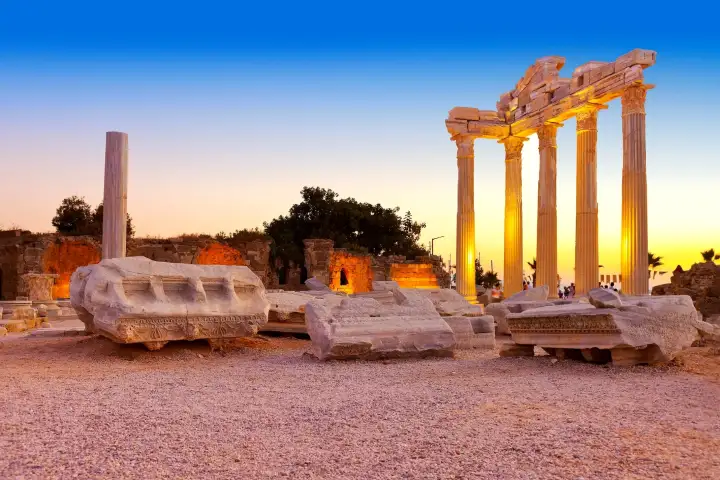 El Templo de Apolo en Side, Antalya, está iluminado con una cálida luz dorada al anochecer, con sus cuatro altas columnas corintias y el entablamento conservado elevándose sobre sarcófagos volcados y bloques de piedra tallada dispersos por el yacimiento antiguo, con un horizonte ámbar brillante detr
