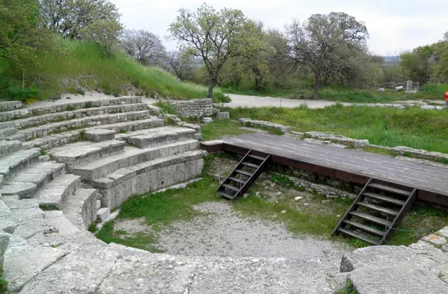 File curvilinee di sedute in pietra calcarea dell'antico teatro di Troia nel sito archeologico di Çanakkale, Turchia, con una pedana moderna in legno accanto alla fossa dell'orchestra e alberi che incorniciano il palcoscenico sulla collina.