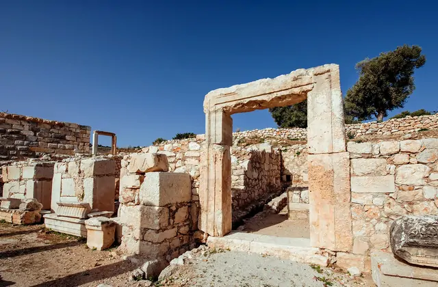 A tall freestanding marble doorway with a massive stone lintel stands amid the ancient ruins of Troy, Çanakkale, Turkey, with low rubble walls stretching behind it under a vivid blue sky.