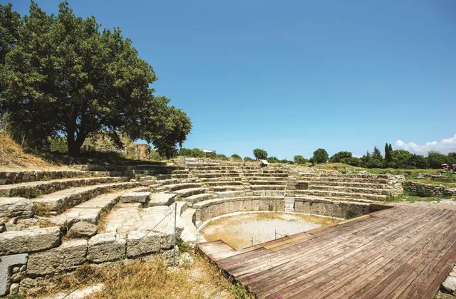 L'odeon ben conservato dell'antica Troia a Çanakkale, Turchia, mostra sedute in pietra a gradoni che si curvano attorno a un'orchestra circolare sabbiosa, con un palco in legno in primo piano e una grande quercia che ombreggia la cavea superiore.
