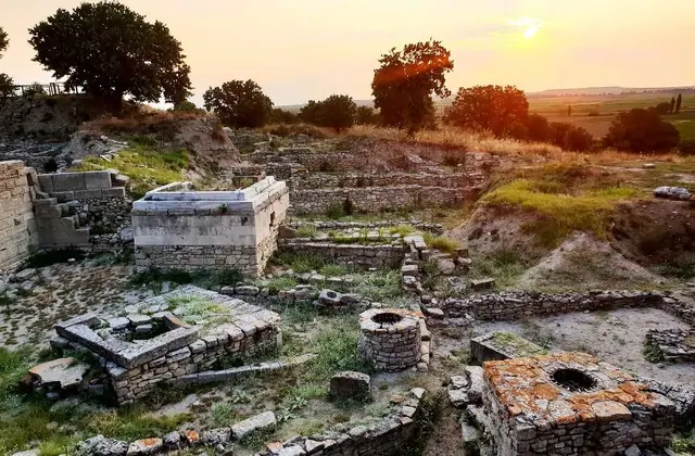 Golden sunset light washes over the multi-room stone foundations of ancient Troy's residential quarter in Çanakkale, Turkey, with circular well openings, a sarcophagus-like marble block, and the flat Troad plain glowing orange on the horizon.