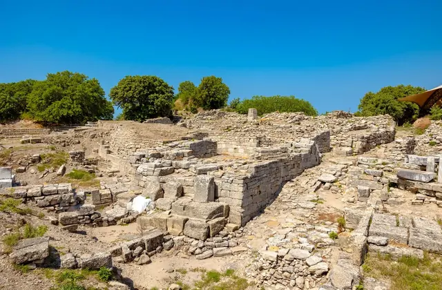 Un amplio panorama del yacimiento arqueológico de Troya en Çanakkale, Turquía, revela densas fundaciones de muros de piedra de múltiples períodos a través de una meseta blanqueada por el sol, con una carpa de sombra protectora sobre una zona clave de excavación visible en la parte superior derecha.