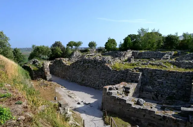 Le massicce mura difensive in pietra a secco dell'antica Troia si ergono lungo uno stretto corridoio scavato nel sito archeologico di Çanakkale in Turchia, con una passerella in legno per i visitatori in fondo e la pianura costiera egea visibile oltre.