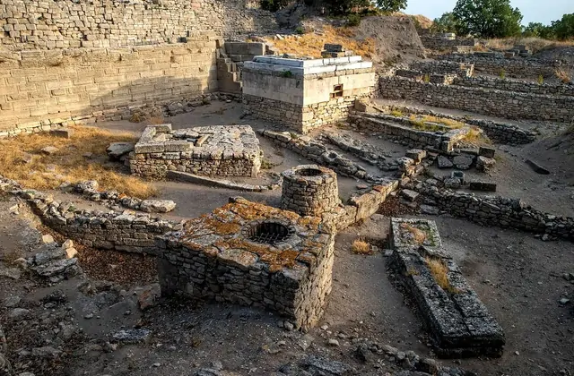 Una vista ravvicinata della zona di scavo stratificata nel sito archeologico di Troia a Çanakkale, Turchia, mostra resti di muri in pietra piccola su più livelli con incavi circolari e una grande base di stele in marmo visibile tra le aride rovine estive.