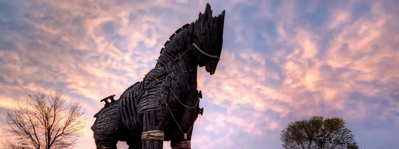 El imponente monumento del Caballo de Troya de madera oscura en el paseo marítimo de Çanakkale en Turquía se recorta en silueta contra un dramático cielo de atardecer rosa y morado, con el puerto de los Dardanelos y los barcos amarrados brillando suavemente al fondo.