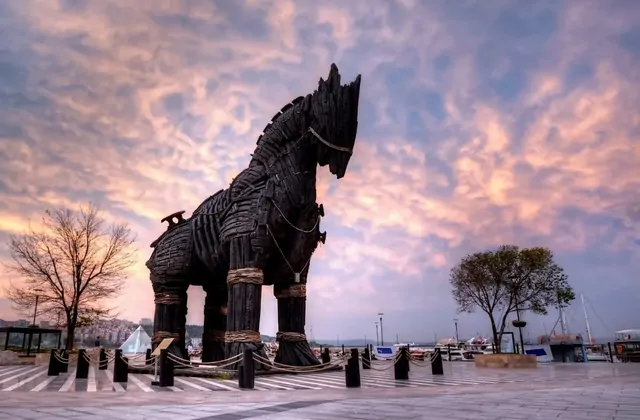 El imponente monumento del Caballo de Troya de madera oscura en el paseo marítimo de Çanakkale en Turquía se recorta en silueta contra un dramático cielo de atardecer rosa y morado, con el puerto de los Dardanelos y los barcos amarrados brillando suavemente al fondo.