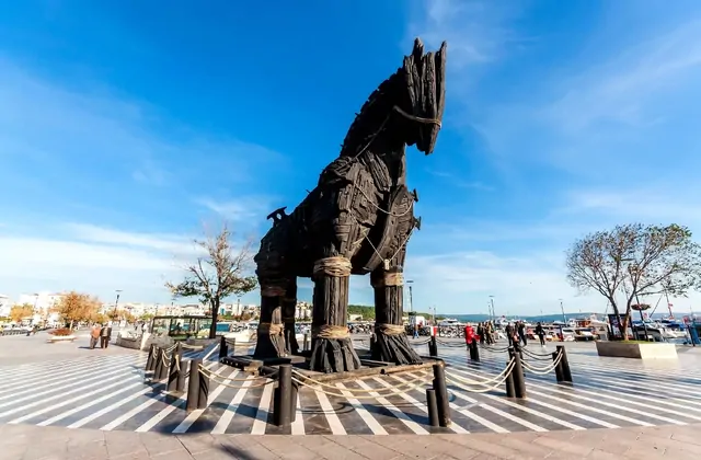 La replica a grandezza naturale del Cavallo di Troia in legno scuro si trova sul lungomare di Çanakkale sotto un cielo blu limpido, con lo stretto dei Dardanelli, barche ormeggiate ed edifici cittadini in lontananza dietro il monumento recintato.