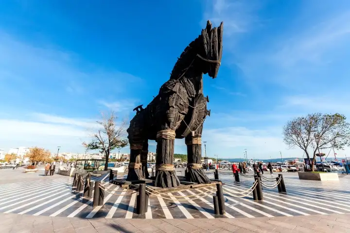 La replica a grandezza naturale del Cavallo di Troia in legno scuro si trova sul lungomare di Çanakkale sotto un cielo blu limpido, con lo stretto dei Dardanelli, barche ormeggiate ed edifici cittadini in lontananza dietro il monumento recintato.