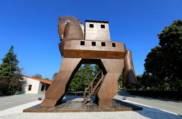 La replica in legno a grandezza naturale del Cavallo di Troia all'ingresso del sito archeologico di Troia a Çanakkale, Turchia, domina il piazzale in legno rossastro levigato, con un corpo a torre rettangolare cavo, finestre aperte e una scala che accede al suo interno.