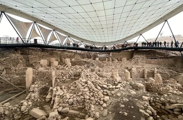 The Göbekli Tepe excavation site viewed under its large triangular tensile canopy roof shows multiple active dig areas with scattered T-shaped pillars and stone walls, while a line of visitors on an elevated walkway silhouetted against the canopy's white fabric look down into the ancient enclosures