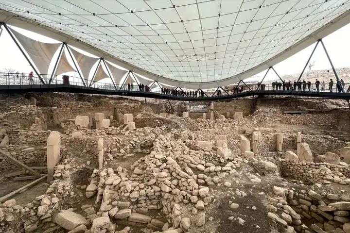 The Göbekli Tepe excavation site viewed under its large triangular tensile canopy roof shows multiple active dig areas with scattered T-shaped pillars and stone walls, while a line of visitors on an elevated walkway silhouetted against the canopy's white fabric look down into the ancient enclosures 