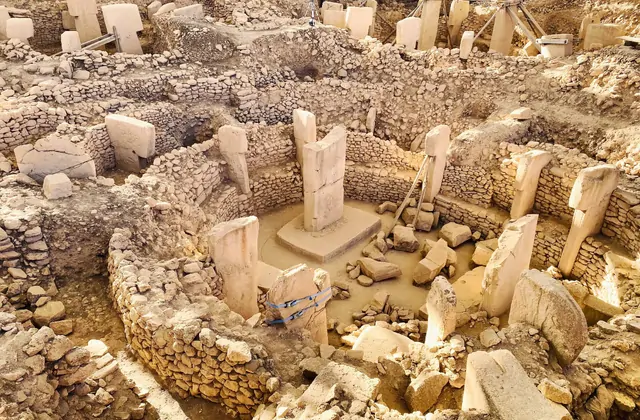 A steep bird's-eye view into Göbekli Tepe's most excavated enclosure reveals a tightly-packed circular chamber with two central T-shaped pillars on a raised stone base, surrounded by at least a dozen peripheral pillars of varying heights set into the curved dry-stone perimeter wall.