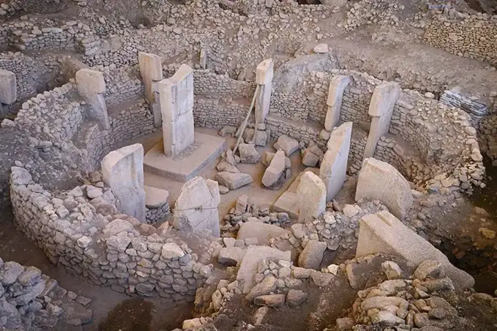 An oblique aerial view of a Göbekli Tepe enclosure in natural daylight shows the full circular arrangement of T-shaped limestone pillars around a central pair, fallen stone slabs littering the sandy floor, and the curved rubble wall clearly defining the enclosure boundary.