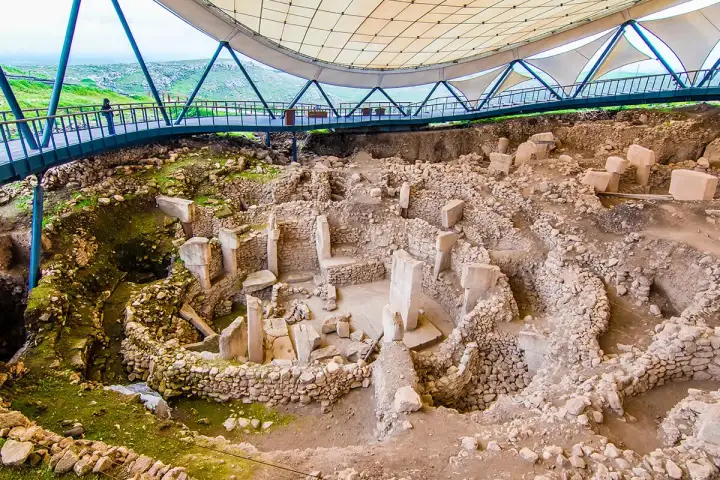 Inside Göbekli Tepe's modern protective canopy on a lush green spring day, a close-angled view shows several T-shaped limestone pillars still partially embedded in excavation soil within a circular stone enclosure, with visitors visible on the steel walkway bridge above and vivid green hills framed 