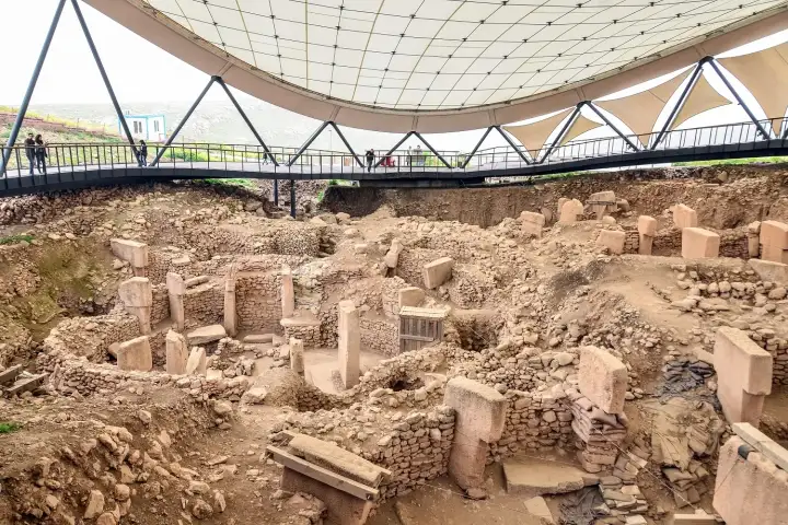 The Göbekli Tepe archaeological site seen from inside the protective canopy structure shows the broad excavation field with rectangular stone-built chambers, multiple upright T-shaped pillars at varying stages of excavation, and two visitors visible on the walkway silhouetted against the light flood