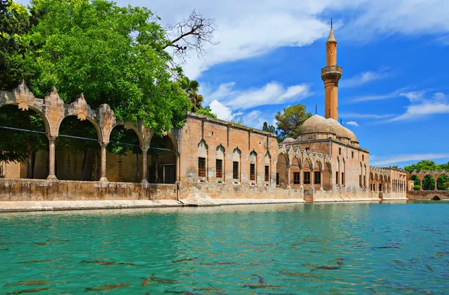 A sunny wide-angle view of Balıklıgöl — the Pool of Abraham — in Şanlıurfa shows the full length of the Halil-ur Rahman Mosque's ornate stone arcaded facade bordering the vivid turquoise pool, its minaret rising above green trees against a bright blue sky, with sacred carp visible beneath the water'