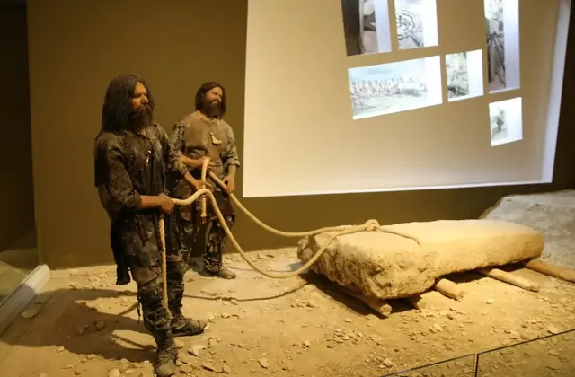 Un diorama a tamaño real en el Museo de Şanlıurfa ilustra el transporte de piedras neolíticas en Göbekli Tepe: dos hombres prehistóricos realísticamente reconstruidos con cabello largo y ropa de cuero arrastran un bloque de piedra caliza rugosa sobre rodillos de madera usando una gruesa cuerda, con